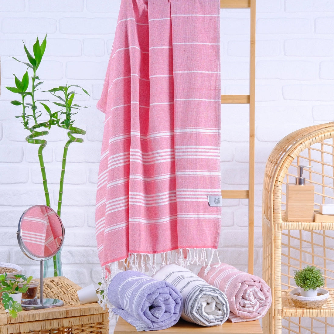 A pink Turkish towel with white stripes is hanging on a wooden rack next to a wicker basket with a green plant and a blue towel. The towel is also displayed on a lower shelf with a white and blue towel.
